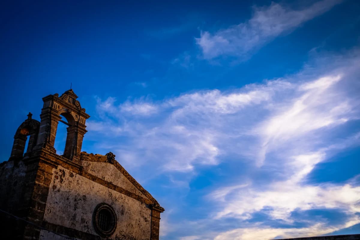 A historic church with a bell tower against a vibrant blue sky with scattered clouds in Marzamemi, Italy.