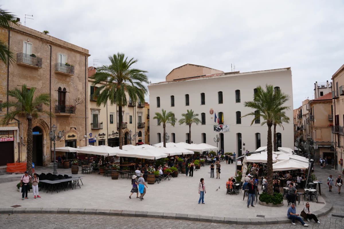 Busy Piazza Del Duomo in Cefalù, Italy, bustling with people and cafes under a cloudy sky.